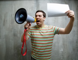 man shouting into a megaphone and holding a white sheet of paper on a background of gray textured wall